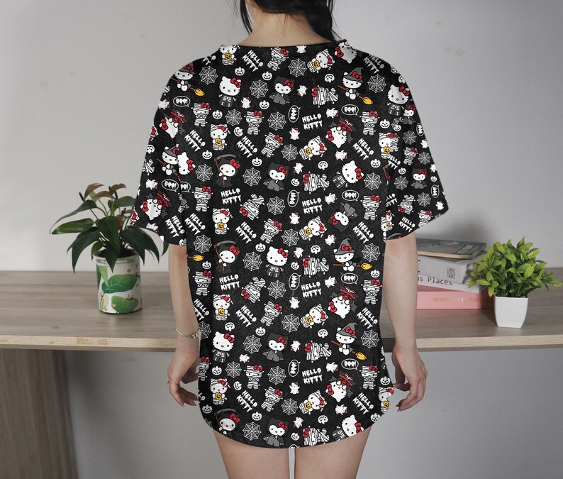 May include: Black short-sleeved shirt with an all-over print featuring Hello Kitty characters, bats, spiderwebs, and the text "Hello Kitty". The design includes white, red, and black elements.