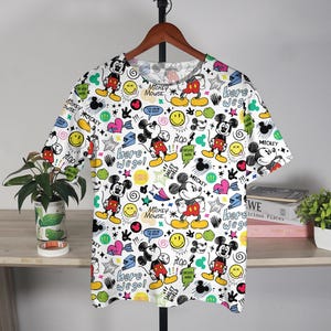 May include: White t-shirt with a repeating pattern of Mickey Mouse, smiley faces, stars, and text. The shirt features Mickey in red shorts and yellow shoes. Text includes "Mickey Mouse" and "Here we go!"