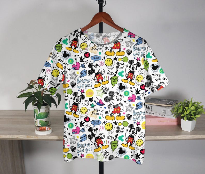 May include: White t-shirt with a repeating pattern of Mickey Mouse, smiley faces, stars, and text. The shirt features Mickey in red shorts and yellow shoes. Text includes "Mickey Mouse" and "Here we go!"