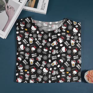 May include: Black V-neck t-shirt with a Halloween-themed Hello Kitty pattern. The design includes Hello Kitty characters dressed as mummies, witches, and vampires, along with spiderwebs and the text "Hello Kitty".