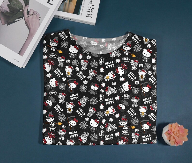 May include: Black V-neck t-shirt with a Halloween-themed Hello Kitty pattern. The design includes Hello Kitty characters dressed as mummies, witches, and vampires, along with spiderwebs and the text "Hello Kitty".