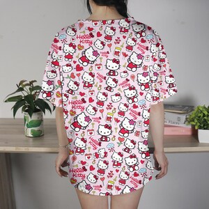 May include: A pink, short-sleeved shirt featuring a repeating pattern of Hello Kitty characters, hearts, and other decorative elements. The design includes the text "Hello Kitty" in red. The shirt is worn by a person.