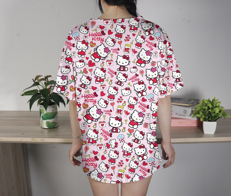 May include: A pink, short-sleeved shirt featuring a repeating pattern of Hello Kitty characters, hearts, and other decorative elements. The design includes the text "Hello Kitty" in red. The shirt is worn by a person.