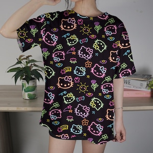May include: Black t-shirt featuring a neon-style pattern of Hello Kitty faces, flowers, stars, and suns. The design includes pink, yellow, green, and orange outlines. The shirt has short sleeves and a round neckline.