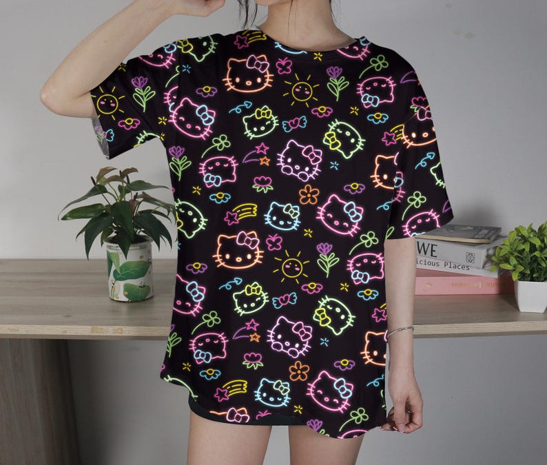May include: Black t-shirt featuring a neon-style pattern of Hello Kitty faces, flowers, stars, and suns. The design includes pink, yellow, green, and orange outlines. The shirt has short sleeves and a round neckline.