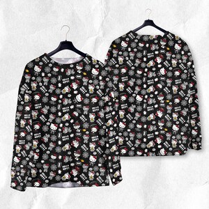 May include: Black long-sleeve shirt with a repeating pattern of Hello Kitty characters, snowflakes, and text. The design includes red, white, and gray elements on a black background. The shirt is displayed on a hanger.