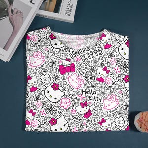 Op de afbeelding: Wit T-shirt met een herhaald patroon van Hello Kitty-personages, bloemen en de tekst "Hello Kitty" in zwart en roze. Het ontwerp bevat roze strikken en bloemmotieven. Het shirt is op een donkerblauwe ondergrond gevouwen.