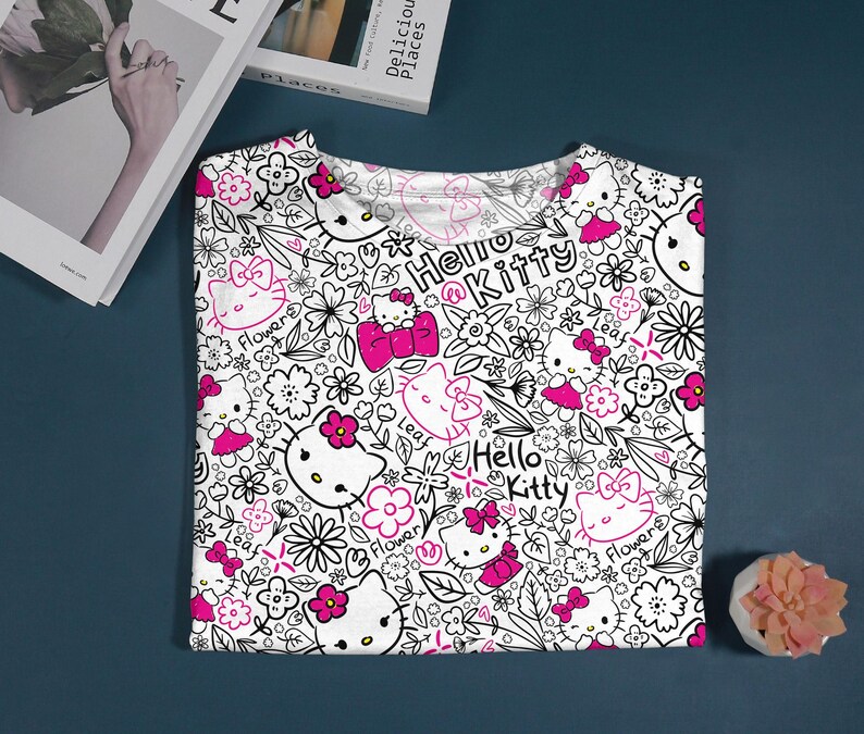 Op de afbeelding: Wit T-shirt met een herhaald patroon van Hello Kitty-personages, bloemen en de tekst "Hello Kitty" in zwart en roze. Het ontwerp bevat roze strikken en bloemmotieven. Het shirt is op een donkerblauwe ondergrond gevouwen.