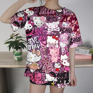 May include: A short-sleeved, oversized t-shirt featuring a vibrant, all-over print of Hello Kitty characters in various poses, interspersed with graffiti-style text in shades of pink and white. The shirt has a crew neck and a loose fit.