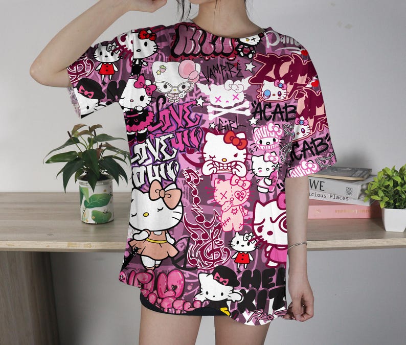 May include: A short-sleeved, oversized t-shirt featuring a vibrant, all-over print of Hello Kitty characters in various poses, interspersed with graffiti-style text in shades of pink and white. The shirt has a crew neck and a loose fit.