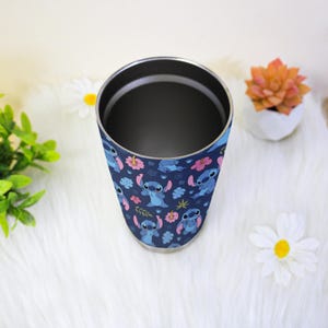 Puede incluir: Un vaso de acero inoxidable con un dise&ntilde;o azul marino que presenta al personaje Stitch de Lilo & Stitch, flores rosas y otros elementos florales. El vaso tiene un borde plateado y un interior oscuro.
