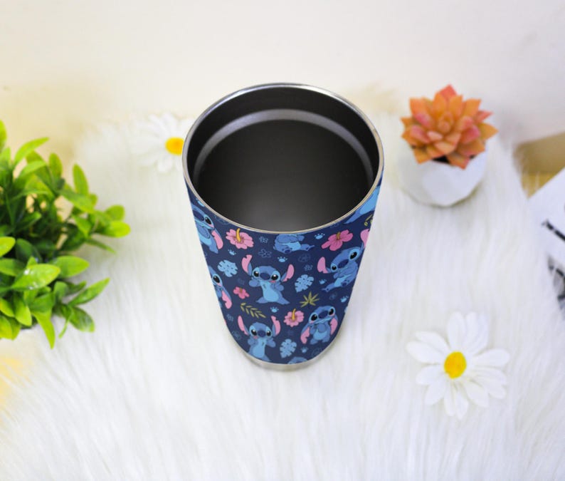 Puede incluir: Un vaso de acero inoxidable con un dise&ntilde;o azul marino que presenta al personaje Stitch de Lilo & Stitch, flores rosas y otros elementos florales. El vaso tiene un borde plateado y un interior oscuro.