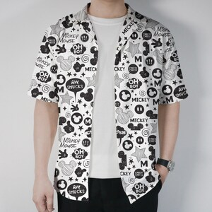 Puede incluir: Camisa blanca de manga corta con un estampado negro de Mickey Mouse. La camisa presenta ilustraciones de Mickey Mouse, estrellas y textos como "Oh Boy" y "Aw Shucks". La camisa está abierta, revelando una camiseta blanca debajo.