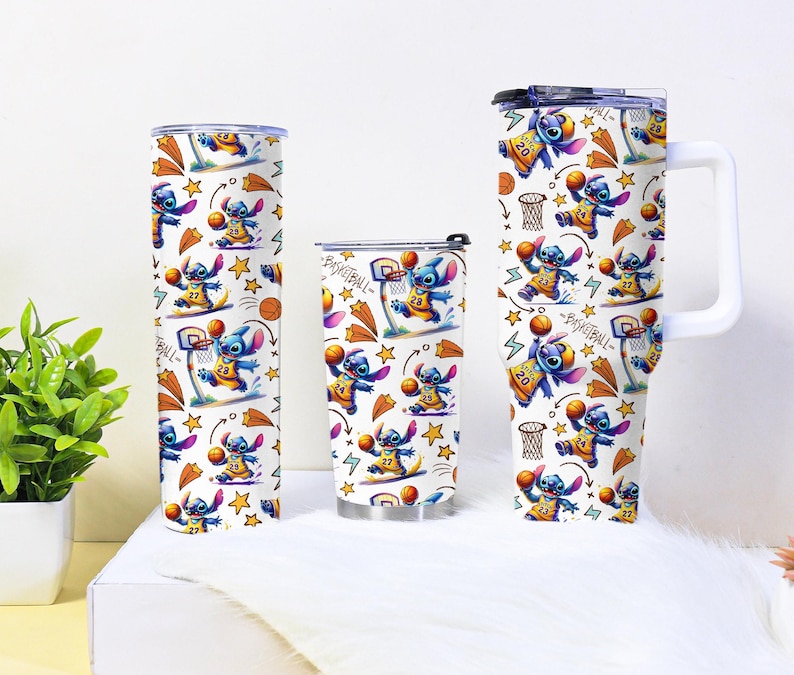 May include: Three white tumblers with a basketball-themed design featuring a blue cartoon character. The design includes basketballs, hoops, and stars. One tumbler is tall and cylindrical, another is shorter, and the third has a handle.