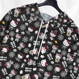 May include: Black hoodie featuring a repeating pattern of Hello Kitty characters in Halloween costumes, spiderwebs, pumpkins, and the text "Hello Kitty". The hoodie has a drawstring and is hanging on a black hanger.