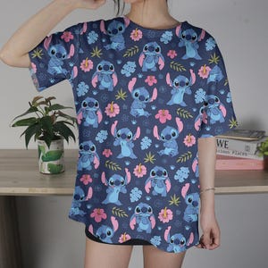 Pu&ograve; includere: Una maglietta blu navy con un motivo ripetuto del personaggio Stitch di Lilo & Stitch. Il design include Stitch, fiori rosa e foglie verdi.
