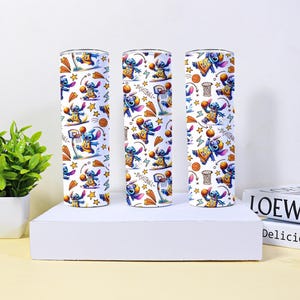 May include: Three white insulated tumblers with a repeating pattern of the character Stitch playing basketball. The design includes basketball hoops, stars, and the word "Basketball". The tumblers are cylindrical and sit on a white platform.