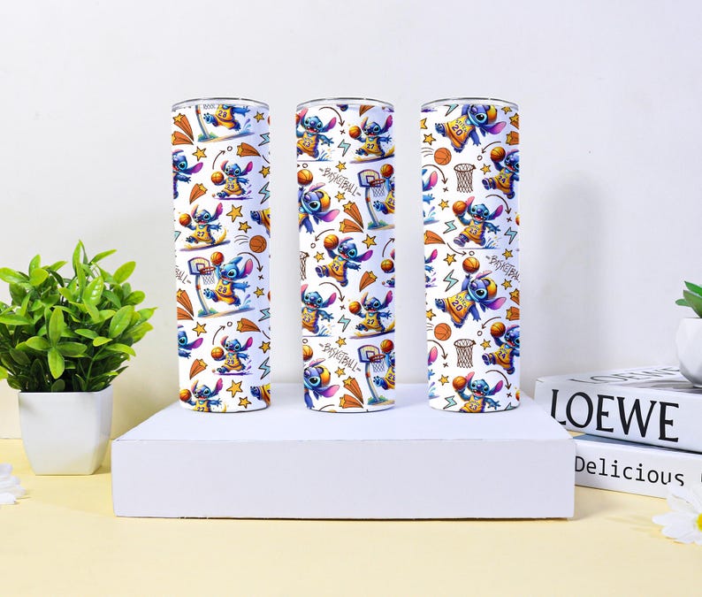 May include: Three white insulated tumblers with a repeating pattern of the character Stitch playing basketball. The design includes basketball hoops, stars, and the word "Basketball". The tumblers are cylindrical and sit on a white platform.