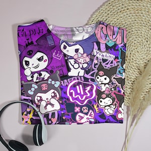 May include: A purple t-shirt with a vibrant graphic design featuring cartoon characters and graffiti-style text. The design incorporates characters in white, black, and purple, with the word "RAFFITI" visible. White and black headphones are in the foreground.