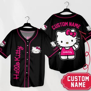 Puede incluir: Camiseta de b&eacute;isbol negra con gr&aacute;ficos de Hello Kitty. La camiseta tiene un ribete rosa, texto de Hello Kitty en el lateral y un gr&aacute;fico de Hello Kitty en la parte delantera. La parte posterior de la camiseta tiene un gran gr&aacute;fico de Hello Kitty y el texto "CUSTOM NAME".