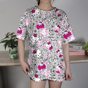 Op de afbeelding: Wit T-shirt met een herhaald patroon van Hello Kitty-gezichten, roze strikken en bloemelementen. De woorden "Hello Kitty" zijn ook in het ontwerp verwerkt. Het shirt heeft korte mouwen en een losse pasvorm.
