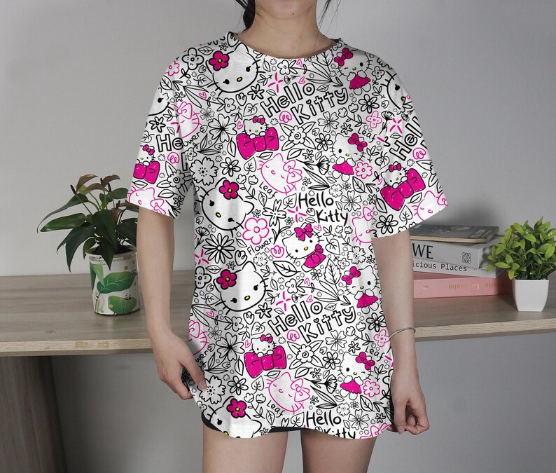 Op de afbeelding: Wit T-shirt met een herhaald patroon van Hello Kitty-gezichten, roze strikken en bloemelementen. De woorden "Hello Kitty" zijn ook in het ontwerp verwerkt. Het shirt heeft korte mouwen en een losse pasvorm.