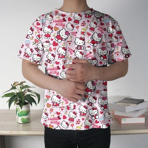 May include: A light pink t-shirt featuring an all-over print of Hello Kitty characters in various poses. The design includes hearts, bows, and the words "Hello Kitty". The shirt has short sleeves and a crew neck.