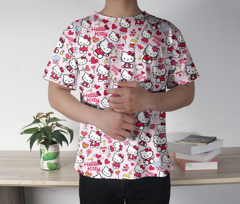 May include: A light pink t-shirt featuring an all-over print of Hello Kitty characters in various poses. The design includes hearts, bows, and the words "Hello Kitty". The shirt has short sleeves and a crew neck.