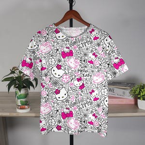 Op de afbeelding: Wit T-shirt met een herhaald patroon van Hello Kitty gezichten, strikken en bloemen in zwart en roze. De woorden "Hello Kitty" staan ook in het ontwerp. Het shirt hangt aan een houten hanger.