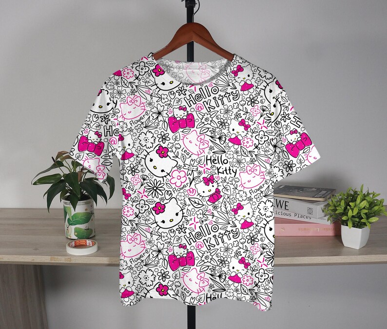 Op de afbeelding: Wit T-shirt met een herhaald patroon van Hello Kitty gezichten, strikken en bloemen in zwart en roze. De woorden "Hello Kitty" staan ook in het ontwerp. Het shirt hangt aan een houten hanger.