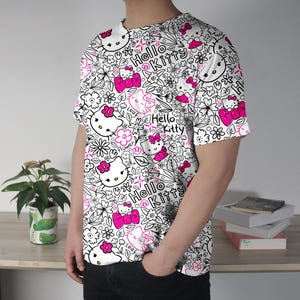 Op de afbeelding: Wit T-shirt met een herhaald patroon van Hello Kitty-personages, bloemen en de woorden "Hello Kitty" in zwart en roze. Het shirt met korte mouwen wordt door een persoon gedragen.
