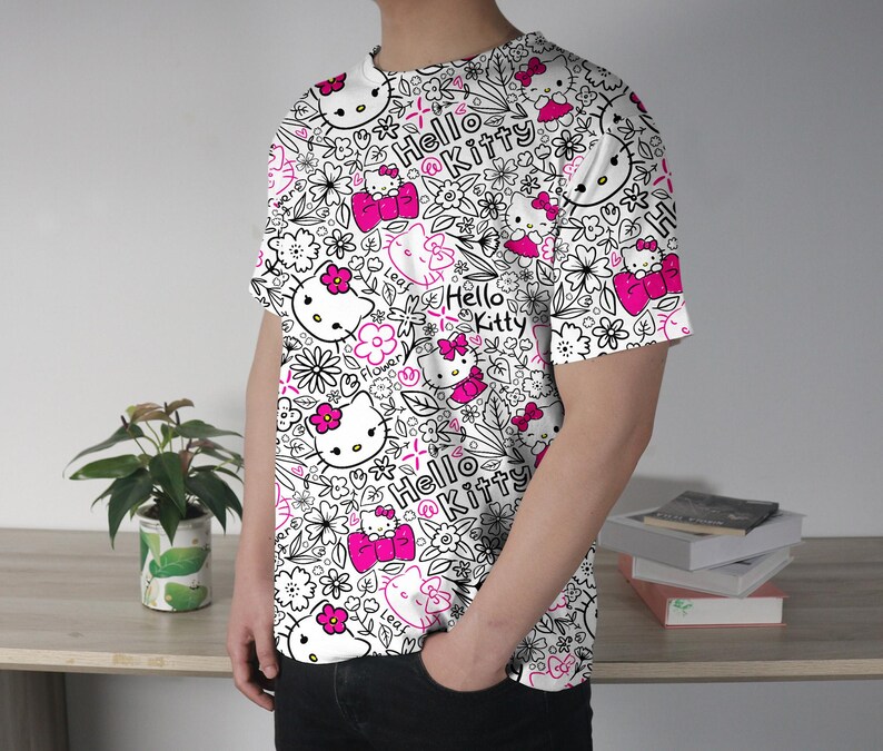 Op de afbeelding: Wit T-shirt met een herhaald patroon van Hello Kitty-personages, bloemen en de woorden "Hello Kitty" in zwart en roze. Het shirt met korte mouwen wordt door een persoon gedragen.