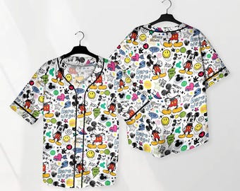 Può includere: Set pigiama bianco con camicia abbottonata e pantaloncini con un colorato motivo di Topolino. Il design include illustrazioni di Topolino, fumetti, stelle, cuori e il testo "Here we go!" e "Mickey Mouse". Rifiniture nere.