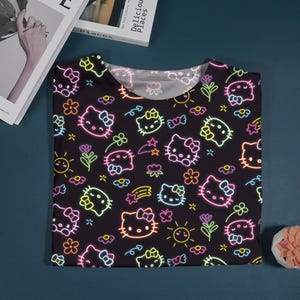 May include: Black t-shirt featuring a neon-style pattern of Hello Kitty faces, flowers, stars, and hearts. The design includes pink, yellow, and green accents. The shirt is folded on a dark blue surface.