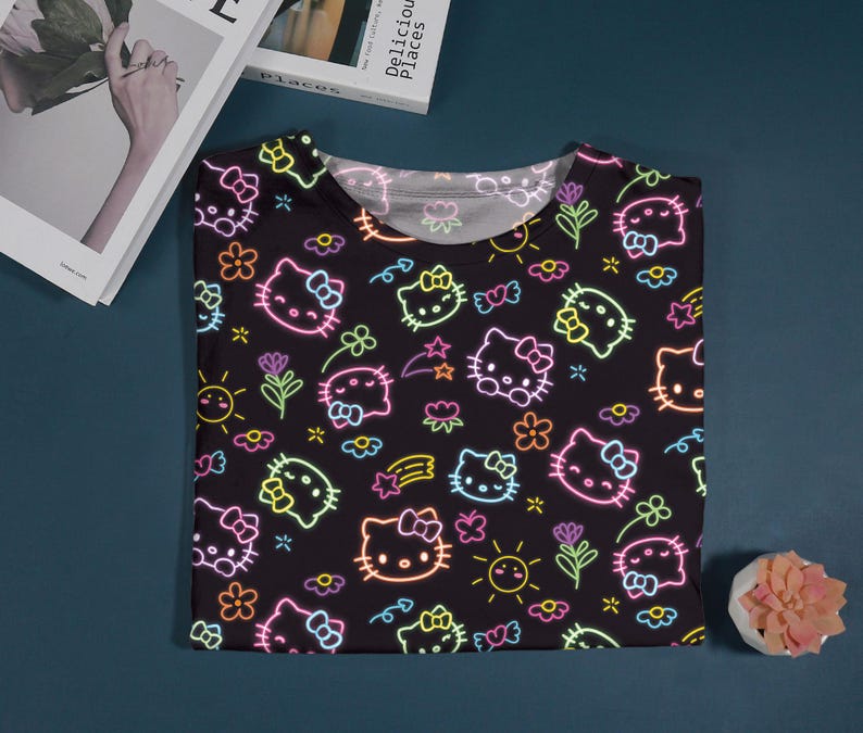 May include: Black t-shirt featuring a neon-style pattern of Hello Kitty faces, flowers, stars, and hearts. The design includes pink, yellow, and green accents. The shirt is folded on a dark blue surface.