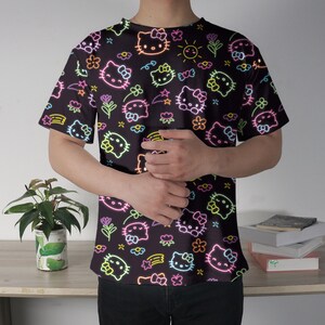 May include: Black t-shirt featuring a neon-style pattern of Hello Kitty characters, flowers, stars, and rainbows. The design is in bright neon colors, creating a vibrant contrast against the dark background. The shirt has short sleeves and a crew neck.
