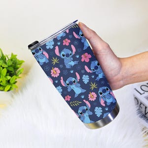 Puede incluir: Un vaso de acero inoxidable con un patr&oacute;n repetido del personaje Stitch de Lilo & Stitch. El dise&ntilde;o incluye a Stitch en varias poses, flores rosas y hojas verdes sobre un fondo azul marino. El vaso tiene una tapa negra.