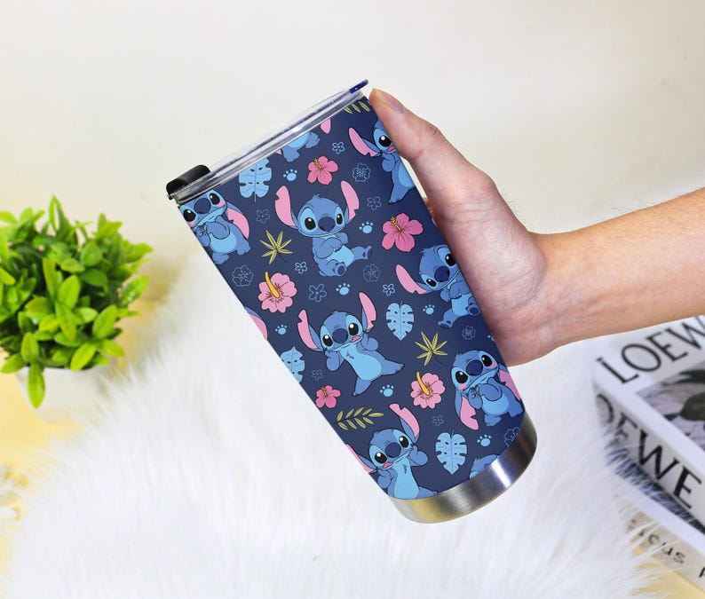 Puede incluir: Un vaso de acero inoxidable con un patr&oacute;n repetido del personaje Stitch de Lilo & Stitch. El dise&ntilde;o incluye a Stitch en varias poses, flores rosas y hojas verdes sobre un fondo azul marino. El vaso tiene una tapa negra.