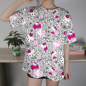 Op de afbeelding: Wit T-shirt met een herhaald patroon van Hello Kitty-personages, bloemen en de woorden "Hello Kitty" in roze. Het ontwerp is zwart omlijnd. Het shirt heeft korte mouwen.