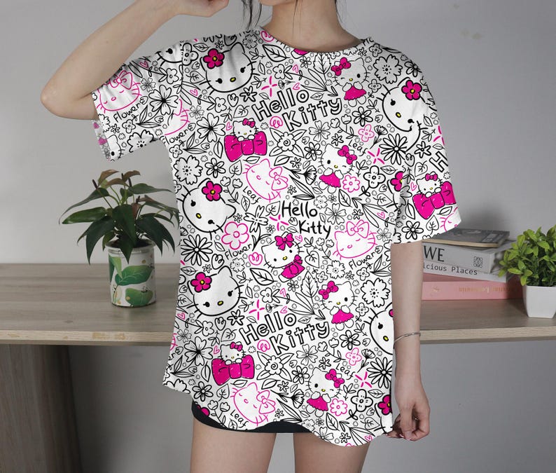 Op de afbeelding: Wit T-shirt met een herhaald patroon van Hello Kitty-personages, bloemen en de woorden "Hello Kitty" in roze. Het ontwerp is zwart omlijnd. Het shirt heeft korte mouwen.