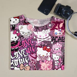 May include: A pink and white t-shirt featuring a repeating pattern of Hello Kitty characters in various poses, with graffiti-style text and skull and crossbones. The shirt has a crew neck and short sleeves.