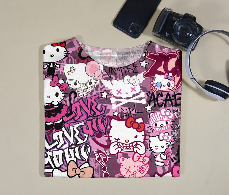 May include: A pink and white t-shirt featuring a repeating pattern of Hello Kitty characters in various poses, with graffiti-style text and skull and crossbones. The shirt has a crew neck and short sleeves.