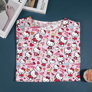 May include: Pink t-shirt featuring a repeating pattern of Hello Kitty characters in various poses, hearts, and other decorative elements. The text "Hello Kitty" is also included in the design. The shirt is folded and displayed on a blue surface.
