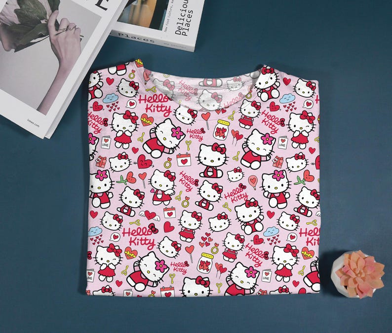 May include: Pink t-shirt featuring a repeating pattern of Hello Kitty characters in various poses, hearts, and other decorative elements. The text "Hello Kitty" is also included in the design. The shirt is folded and displayed on a blue surface.
