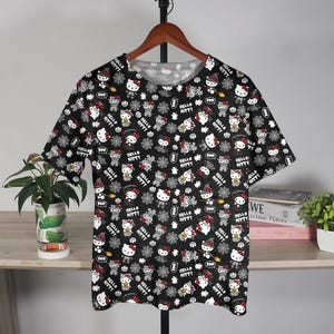May include: Black t-shirt featuring a repeating pattern of Hello Kitty characters in various Halloween costumes. The design includes white, red, and black elements, with the words "Hello Kitty" throughout. The shirt is displayed on a wooden hanger.