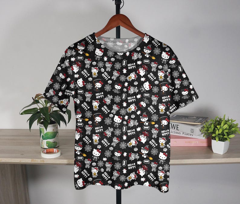 May include: Black t-shirt featuring a repeating pattern of Hello Kitty characters in various Halloween costumes. The design includes white, red, and black elements, with the words "Hello Kitty" throughout. The shirt is displayed on a wooden hanger.