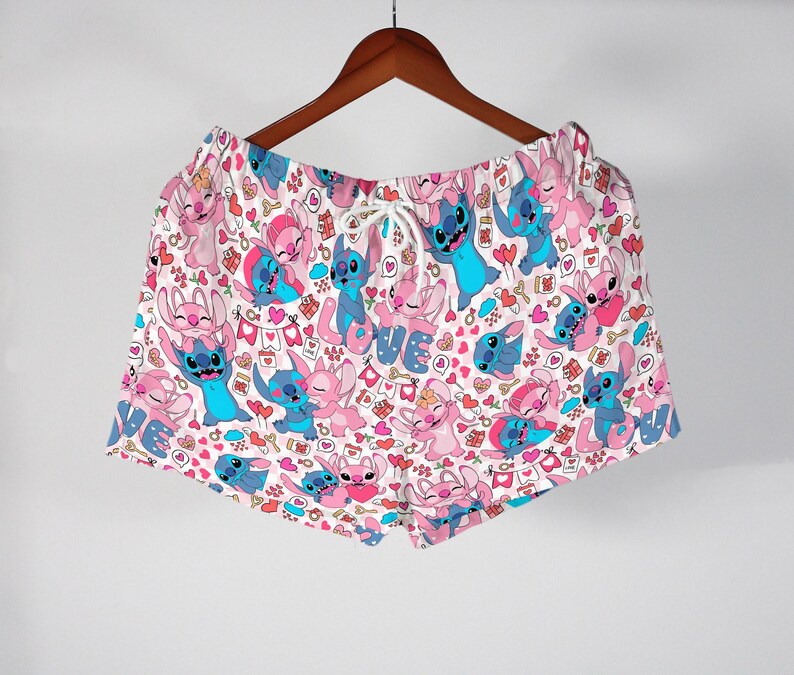 Puede incluir: Shorts blancos con cintura con cord&oacute;n, con un estampado repetido de personajes de dibujos animados azules y rosas, corazones y la palabra "LOVE". Los shorts cuelgan de una percha de madera.