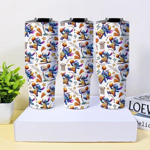 May include: Three white insulated tumblers with black lids. Each tumbler features a repeating pattern of the character Stitch playing basketball, with basketball hoops, stars, and basketballs. The text "Basketball" is also visible.