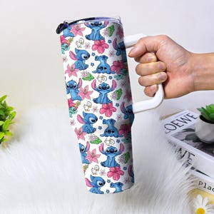 Puede incluir: Un vaso blanco con asa, decorado con un patr&oacute;n repetido de personajes de dibujos animados azules, flores rosas y hojas verdes. El vaso tiene una tapa negra y un asa blanca. El dise&ntilde;o es vibrante y colorido.