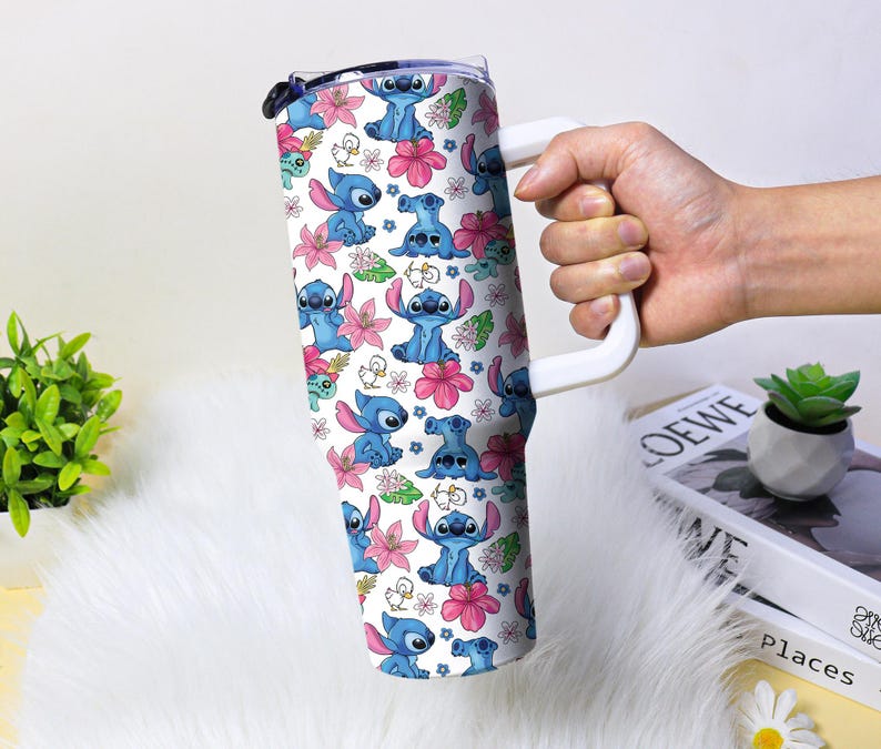 Puede incluir: Un vaso blanco con asa, decorado con un patr&oacute;n repetido de personajes de dibujos animados azules, flores rosas y hojas verdes. El vaso tiene una tapa negra y un asa blanca. El dise&ntilde;o es vibrante y colorido.